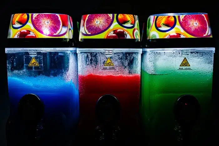 Three illuminated slushi machines with blue, red, and green frozen drinks in transparent tanks against a dark background.