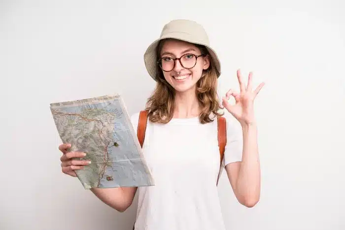 A cheerful female traveler wearing a bucket hat and glasses, holding a map and giving an okay sign against a white background