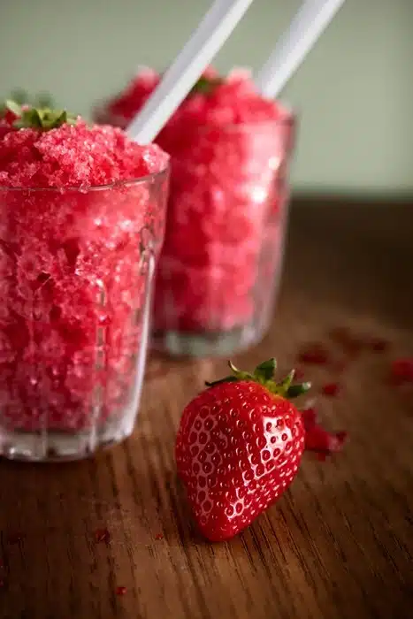 Two glasses of freshly made strawberry slushies with straws on a wooden surface with a fresh strawberry alongside.