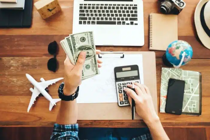 A traveler counting US dollar bills while using a calculator surrounded by travel essentials on a wooden desk