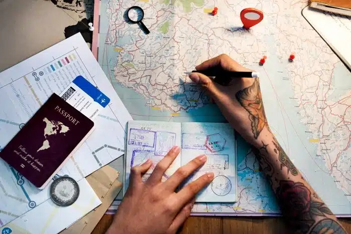 A traveler with tattooed hands planning a trip over a large map, with a passport, boarding pass, and location pins on the table