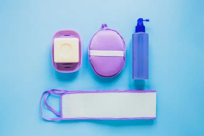 Purple travel bathroom accessories — soap dish, loofah, spray bottle, and back strap laid out on a light blue surface