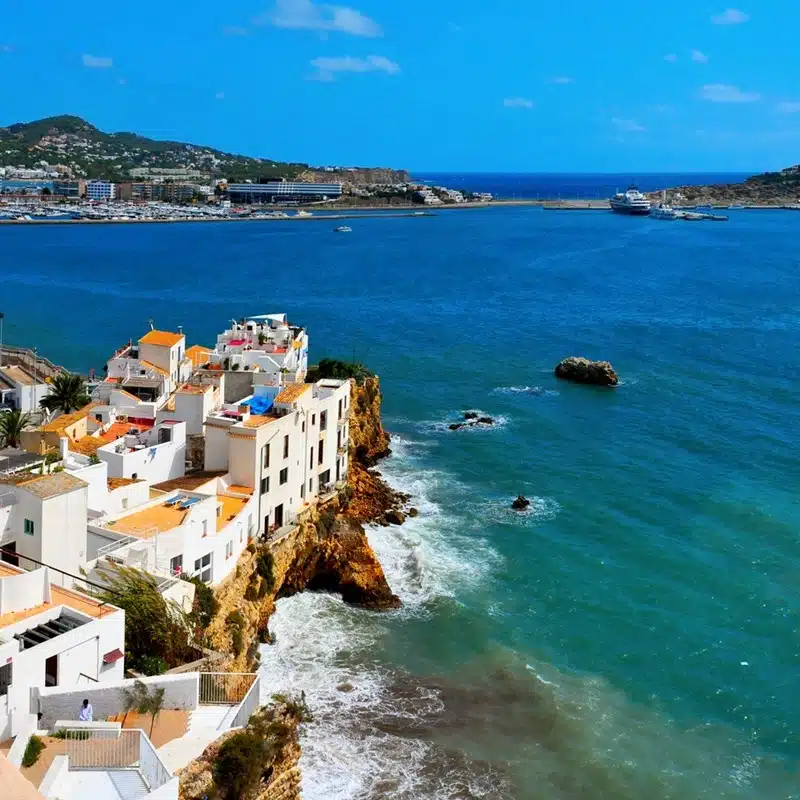 View of Sa Penya district in Ibiza Town with white cliff-top buildings overlooking the blue Mediterranean sea and a ferry in the distance