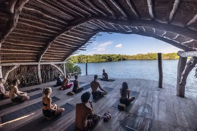 Group meditation session at one of the best wellness retreat destinations, practicing yoga in an open-air thatched pavilion overlooking a tropical lagoon