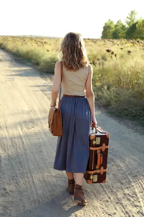 A woman walking alone down a rural dirt road with a vintage suitcase and shoulder bag to maximize solo travel