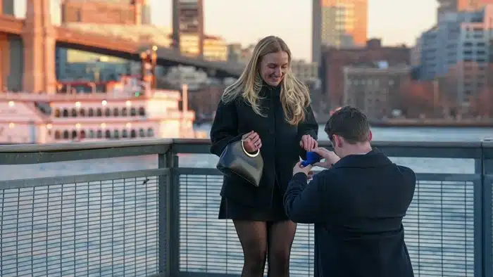 ring for a destination proposal — man kneeling on a waterfront bridge at golden hour with a city skyline in the background