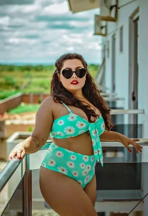 Plus size woman wearing a teal daisy print bikini set with a front tie top and high waist bottoms, posing confidently on a balcony — how to get a summer body on your own terms