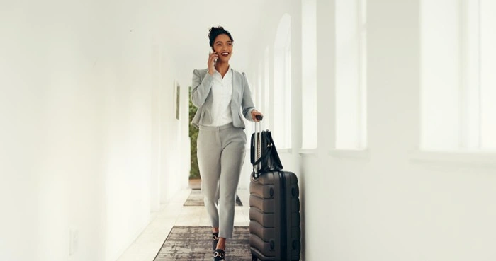 Smiling businesswoman walking through a hotel corridor while on a phone call, pulling a suitcase during a business travel trip
