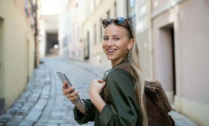 Smiling young woman with a backpack and phone exploring a narrow European cobblestone alley