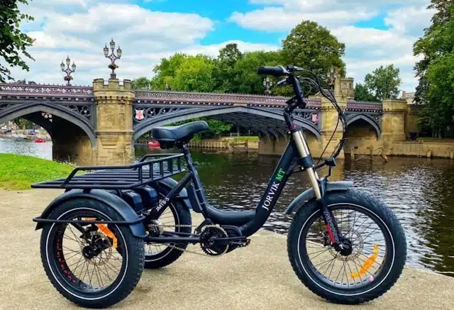 black adult electric tricycle parked by a river with a stone bridge in the background