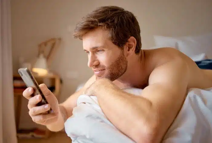 man scrolling through a dating app on his phone in bed