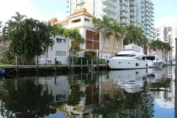 A large yacht docked at a private boat slip beside a vacation house along a Florida canal with palm trees and high-rise buildings