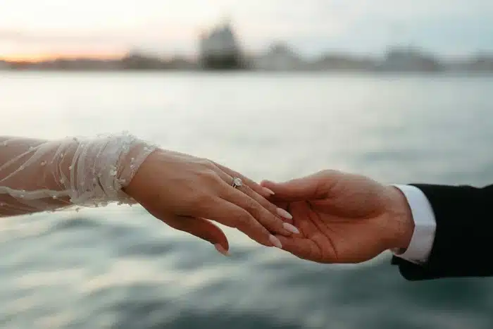 couple holding hands at sunset showing an engagement ring against a soft waterfront backdrop