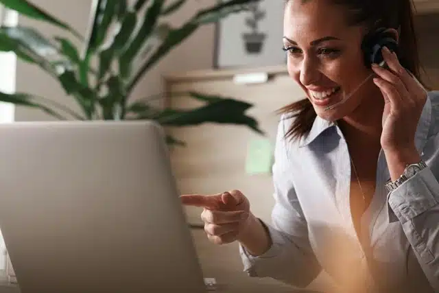 smiling customer service representative wearing a headset while pointing at a laptop screen