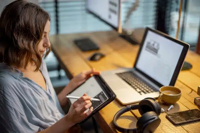 female digital nomad working remotely on a tablet with a laptop and coffee cup on a wooden desk