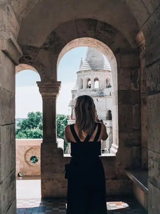 A woman standing under a stone arch at Fisherman's Bastion in Budapest, looking out toward the white turrets