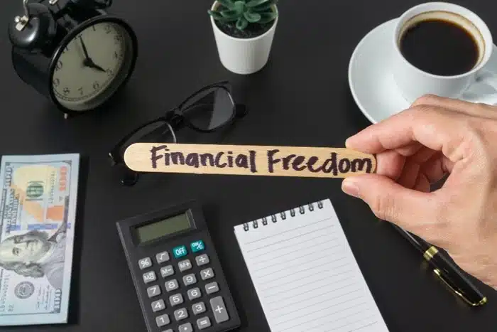 hand holding a financial freedom wooden stick next to a calculator, cash, notebook, and coffee on a dark desk