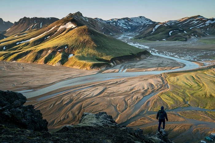Solo hiker standing on a rocky ridge overlooking a vast Icelandic highland landscape with winding rivers and colorful mountains
