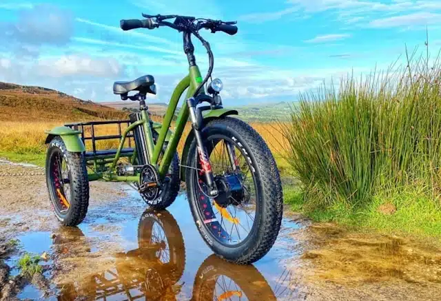 green electric adult tricycle with rear basket on a wet trail surrounded by open countryside