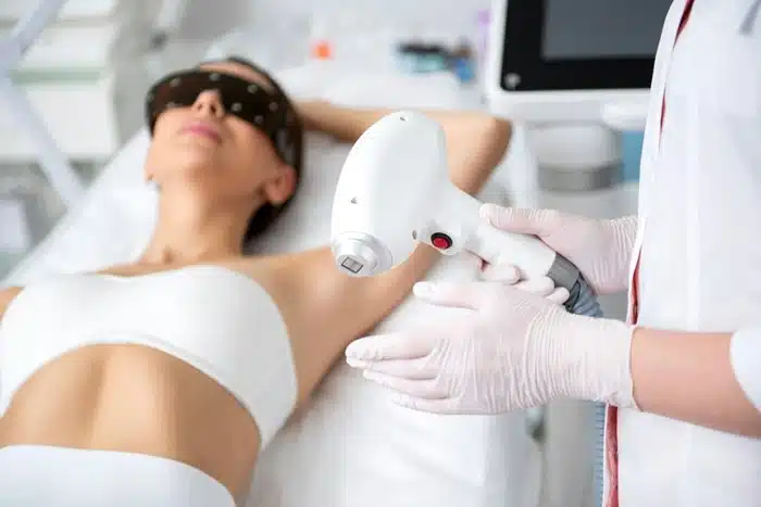 Woman lying on a treatment bed wearing a sleep mask while a technician performs laser hair removal on her underarm area