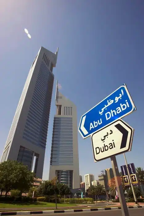 Road sign pointing to Dubai and Abu Dhabi with the Emirates Towers in the background under a clear blue sky