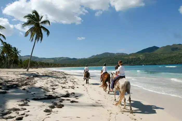 People riding horses along the white sand shore of Playa Rincón in the Dominican Republic with green mountains in the background