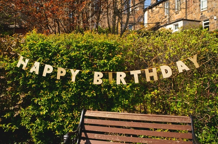 A gold Happy Birthday banner strung across a green garden hedge — the perfect occasion to plan a trip for big life milestones