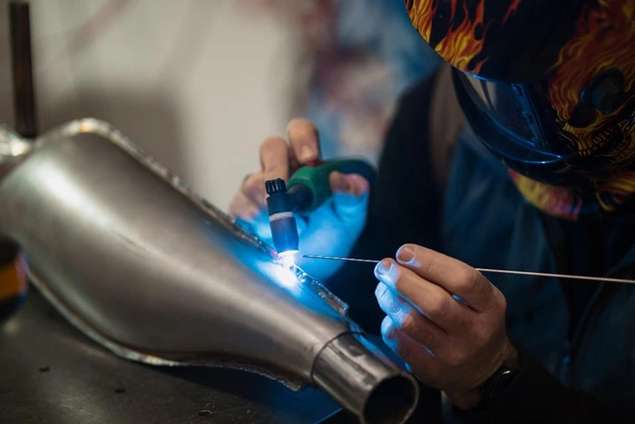welder in a decorative helmet using a plasma cutter on a curved metal piece in a workshop