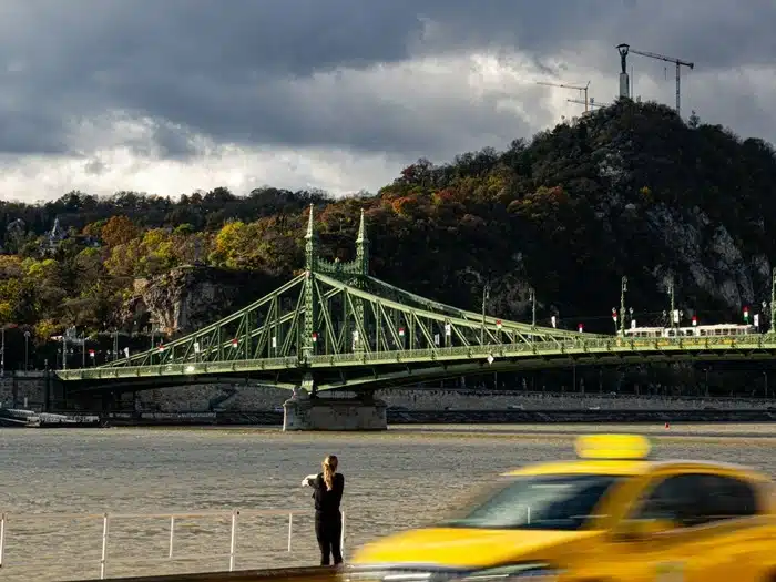 A solo female traveler in Hungary standing by the Danube River facing Szabadság Bridge in Budapest on a cloudy autumn day