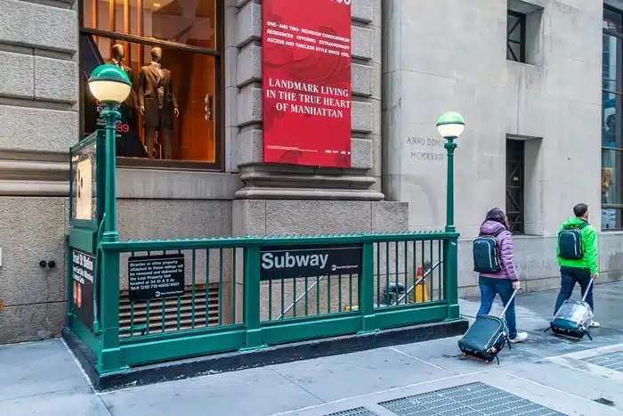 Two tourists with luggage and backpacks walking past a New York City subway entrance in Manhattan