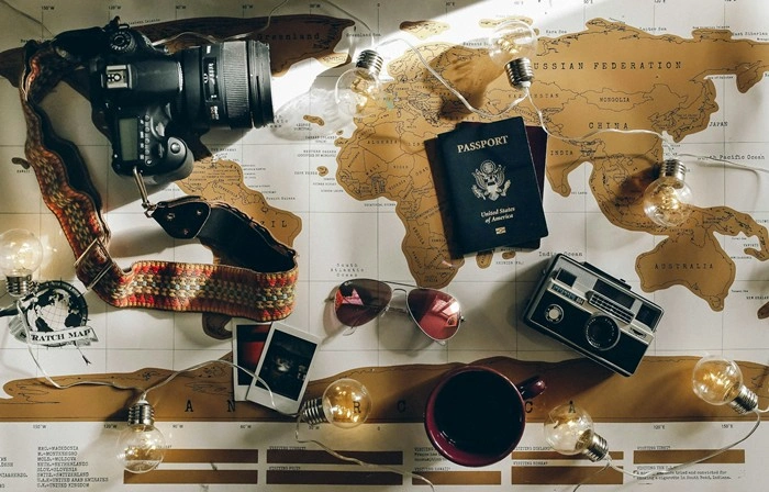 A flat lay of travel essentials including a passport, cameras, sunglasses, and polaroids arranged on a world map