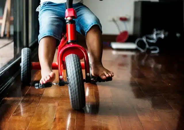young child riding a red kids tricycle barefoot on a wooden floor indoors