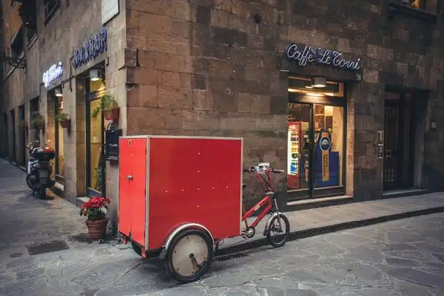 red cargo tricycle with a large box parked on a cobblestone street outside a café