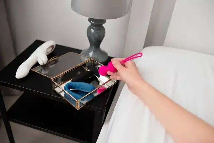 Hand picking up a pink kegel ball from a bedside table with various sex toys on display