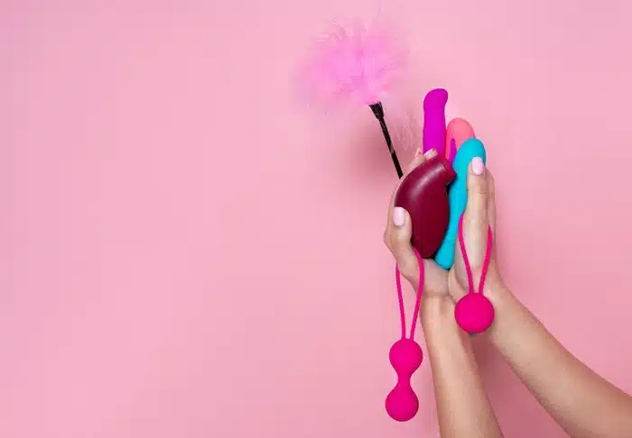 Hands holding a collection of colorful vibrators and kegel balls against a pink background