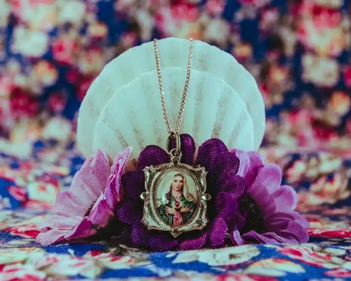 gold mary pendant necklace with colorful portrait displayed on a seashell and purple flowers