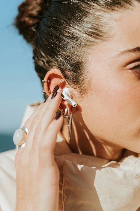 Woman putting on a silver sculptural stud earring with ear cuff and statement ring against a blue sky background