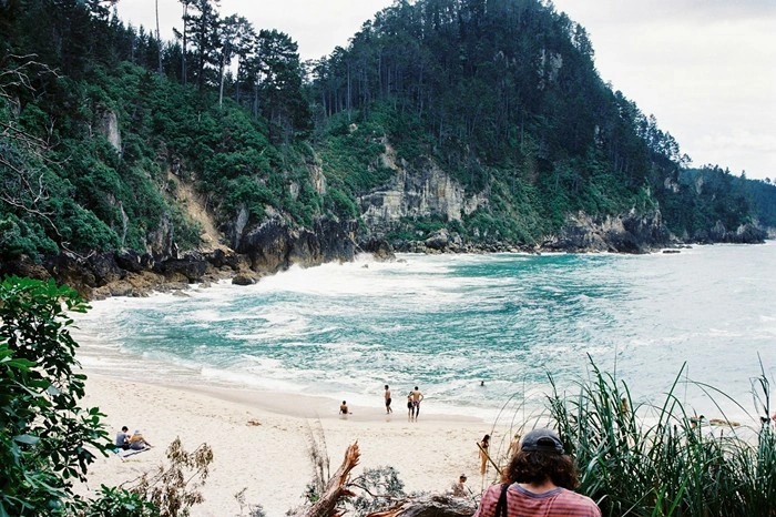 Scenic Whangamata beach in New Zealand with turquoise waves, white sand, and lush forested cliffs in the background