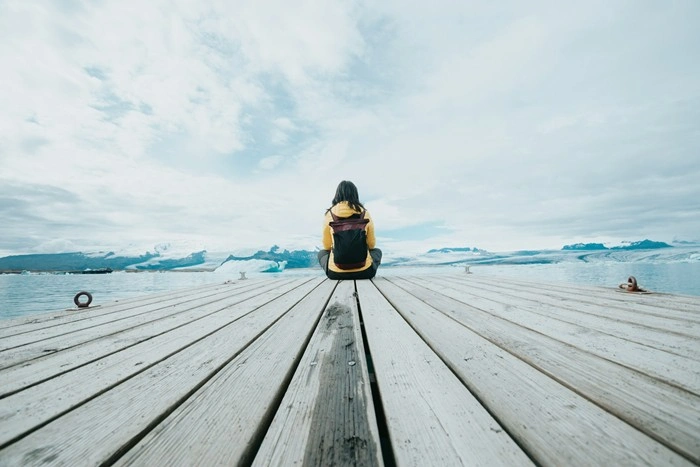 A solo female traveler sitting on a wooden dock overlooking a glacial lagoon with icebergs and mountains in the distance