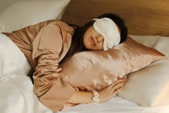 Woman sleeping peacefully on a satin pillow wearing a silk eye mask, representing the importance of quality rest for body health and recovery