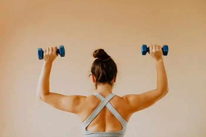 Woman seen from behind lifting blue dumbbells overhead in a grey sports bra, working out as part of a consistent fitness routine
