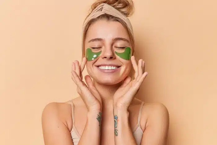 smiling woman with clear healthy skin applying green under-eye patches as part of her skincare routine