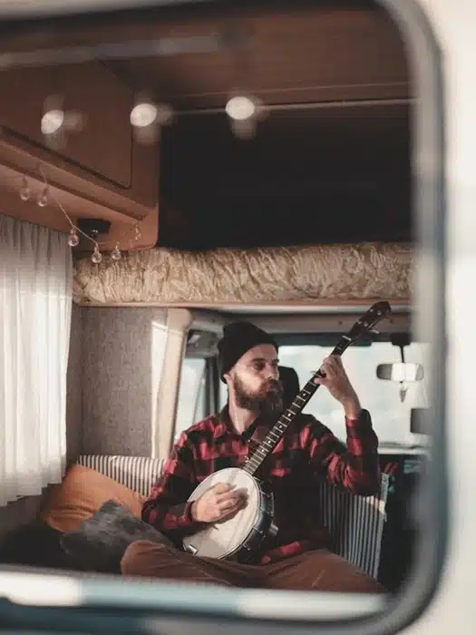 man with a beanie playing banjo on a motorhome bed reflected through a window with fairy lights overhead