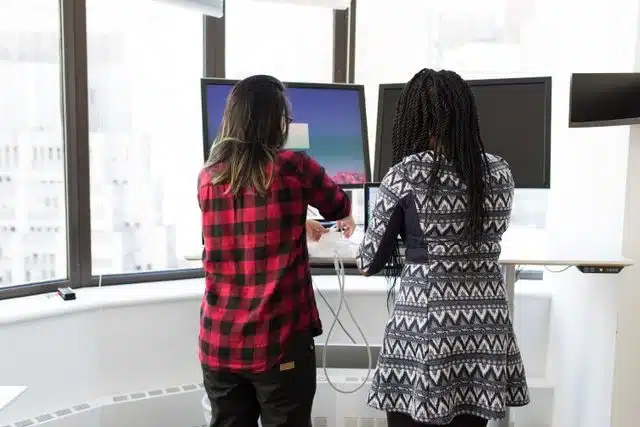 Two students setting up dual monitors on a budget-friendly standing desk by a bright window