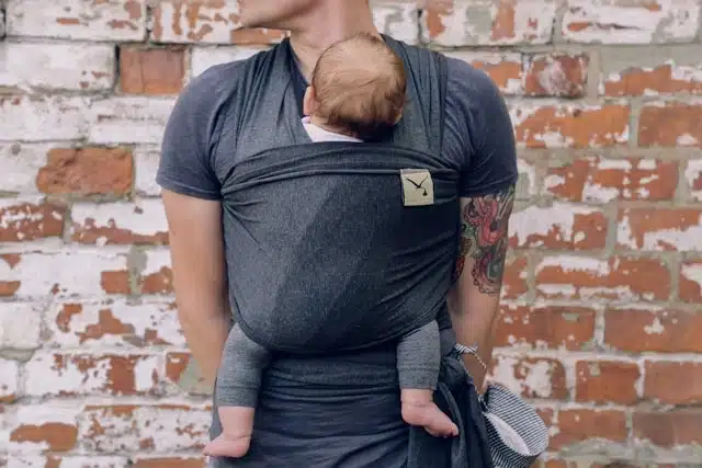 Dad carrying his baby in a wrap carrier while standing against a brick wall