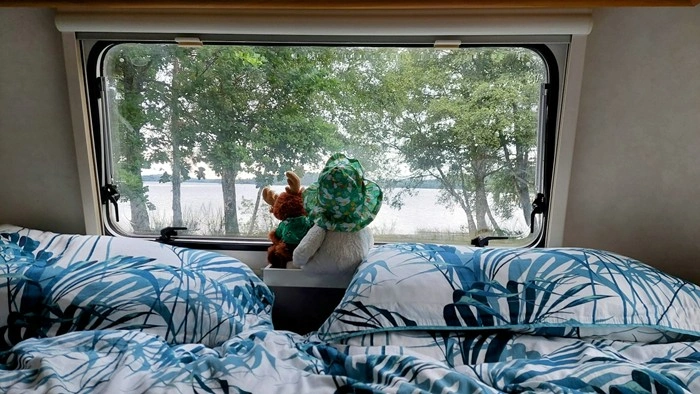 A teddy bear sitting on an RV bed looking out the window at a lakeside view surrounded by trees