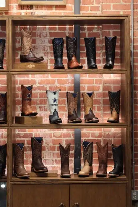 A display of western cowboy boots in various styles and leather finishes arranged on wooden shelves against a brick wall in a Texas boot store