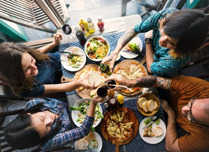 Group of friends toasting drinks over a shared travel meal with pizza, salads, and local dishes on the table