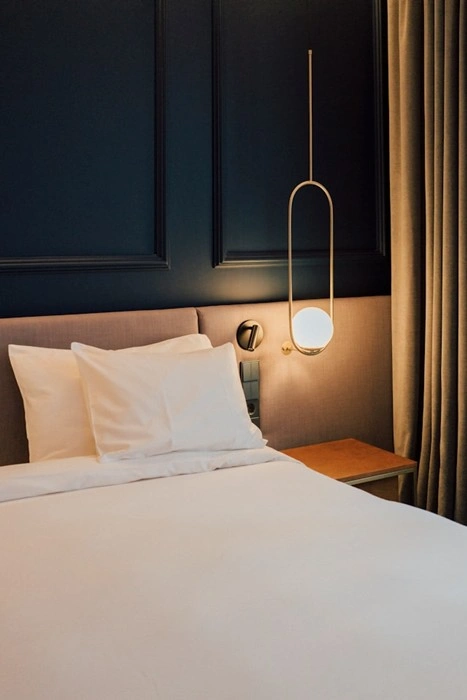 A dark, calm hotel bedroom with a glowing bedside pendant lamp, white crisp pillows, and thick curtains — the kind of controlled darkness that helps you sleep deeply