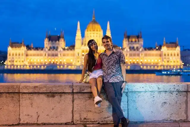 A couple posing by the Danube River with the illuminated Hungarian Parliament Building in Budapest at night